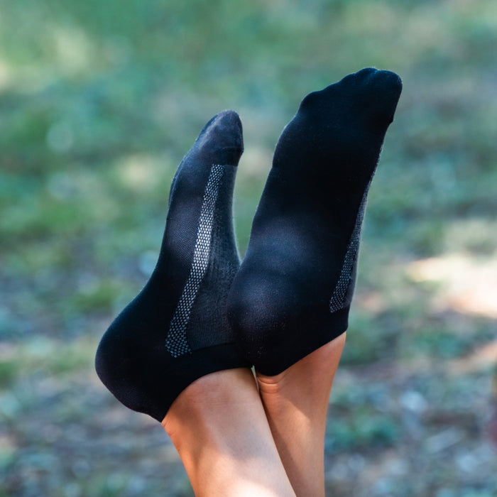 Close-up of a person’s feet wearing black ankle socks with gray stripes, raised and crossed at the ankles, set against a blurred outdoor background with greenery.